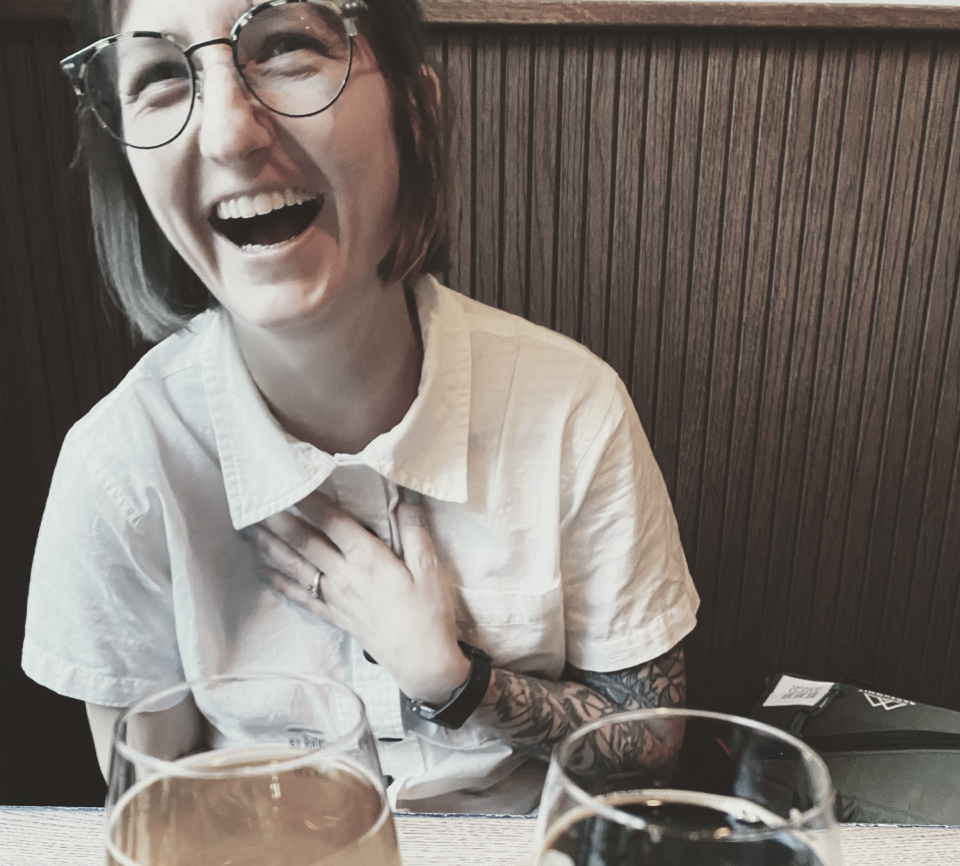 Caroline, wearing eyeglasses and a white collared shirt is laughing joyfully with hand to chest, mouth open. In front of her sits two beers in glasses, one light and one dark.